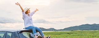 Woman sitting on a car with arms raised in celebration, looking at beautiful mountain scenery.