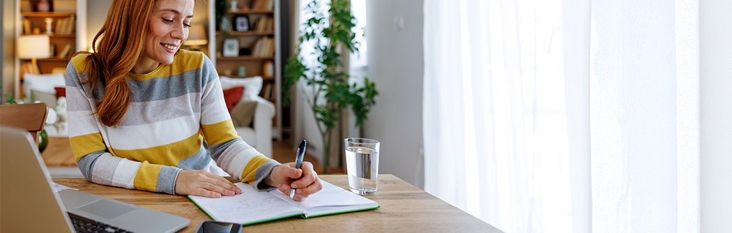 Woman seated at a dining table, planning her time with a notebook and laptop.