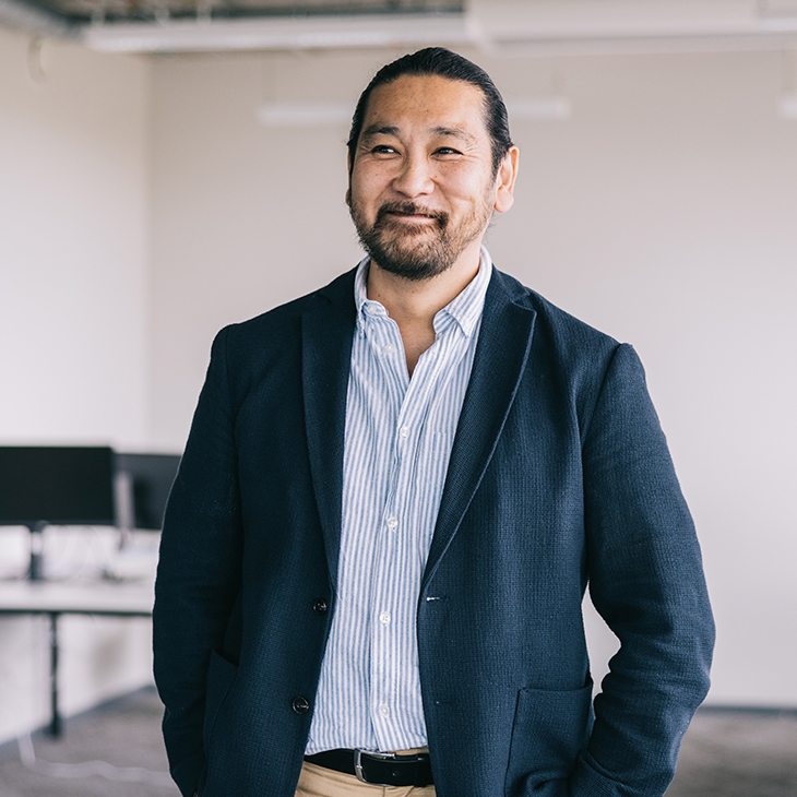 A smiling professional man in a navy blazer stands in a modern, open office.