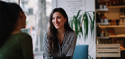 A woman in a striped shirt smiles while holding a tablet and talking to a colleague in a bright, modern cafe setting.