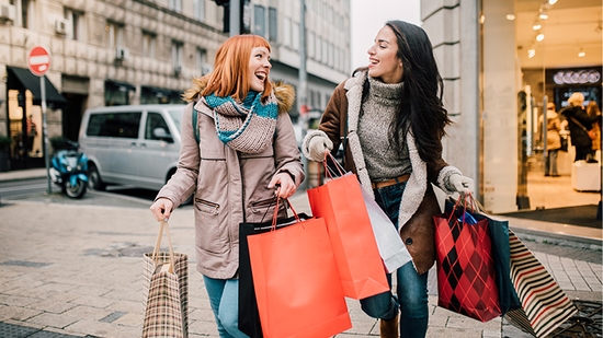 Two smiling women in winter clothes walk down a city street carrying several shopping bags.