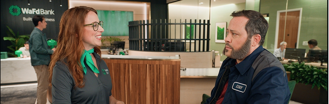 A smiling WaFd Bank employee wearing a green scarf talks to a male customer in a work jacket inside a WaFd branch.