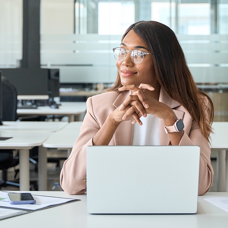 Smiling woman wearing glasses, sitting at an office desk with a laptop and papers.