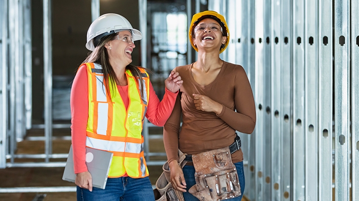 Two women in hard hats and safety gear laugh together while walking through a building under construction with metal wall framing.