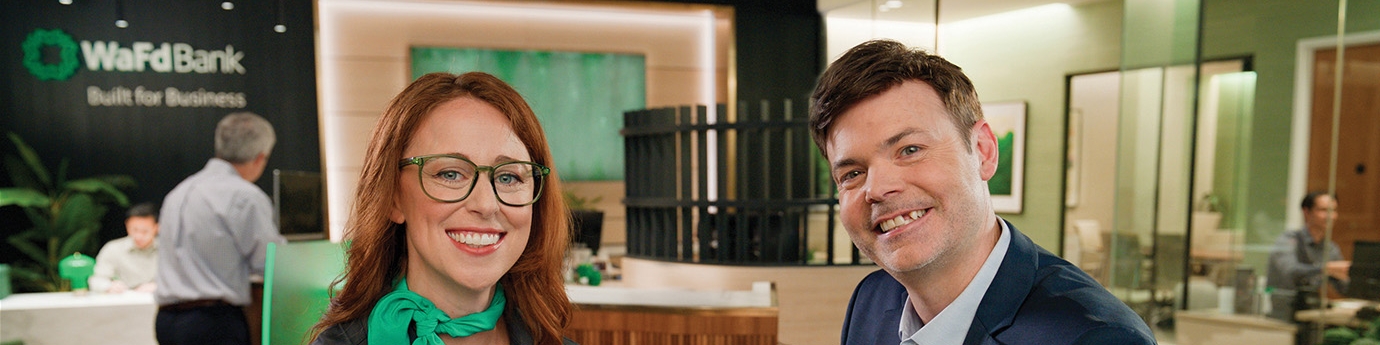 A smiling woman in a WaFd Bank uniform and a man in a blue suit sitting together inside a modern WaFd Bank branch.