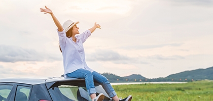 Woman sitting on a car with arms raised in celebration, looking at beautiful mountain scenery.