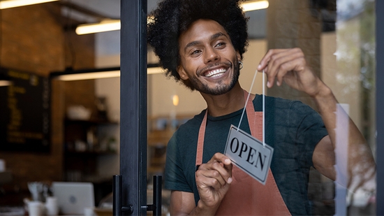 A smiling man with an afro hangs an "OPEN" sign on the glass door of his business.