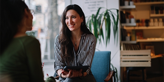 A woman in a striped shirt smiles while holding a tablet and talking to a colleague in a bright, modern cafe setting.