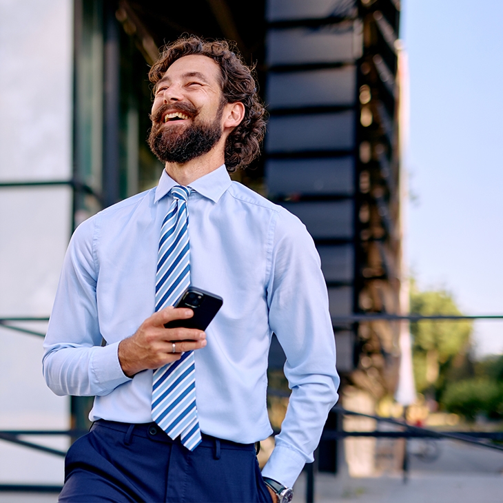 A bearded man in a light blue shirt and striped tie is standing outdoors in front of a modern glass building, laughing while holding a smartphone.