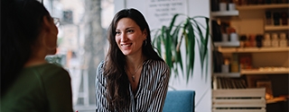 A woman in a striped shirt smiles while holding a tablet and talking to a colleague in a bright, modern cafe setting.