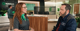 A smiling WaFd Bank employee wearing a green scarf talks to a male customer in a work jacket inside a WaFd branch.