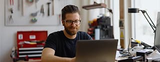 Man with glasses and a beard smiling while working on a laptop at a workbench in a workshop.