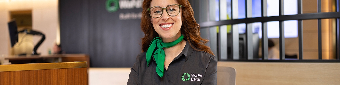 A smiling WaFd Bank representative in a branded shirt and green scarf sits at a desk, providing professional support to business clients.