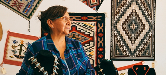 An older woman in a plaid shirt stands in a shop featuring Native American jewelry and woven rugs on the wall.