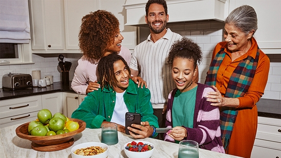 A multi-generational family gathered in a kitchen, smiling as a teenage boy holds a smartphone and a young girl examines a debit card.