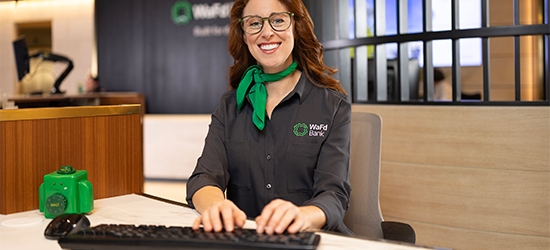 A smiling WaFd Bank representative in a branded shirt and green scarf sits at a desk, providing professional support to business clients.