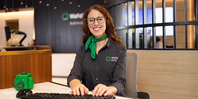 A smiling WaFd Bank representative in a branded shirt and green scarf sits at a desk, providing professional support to business clients.