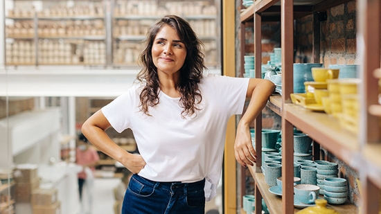 A smiling woman in a white t-shirt leans against wooden shelves filled with handmade ceramic pottery in a bright studio.