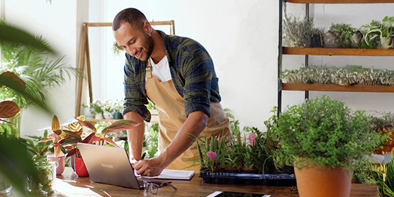 A business owner in an apron working on a laptop at a wooden table in a plant-filled shop or nursery.