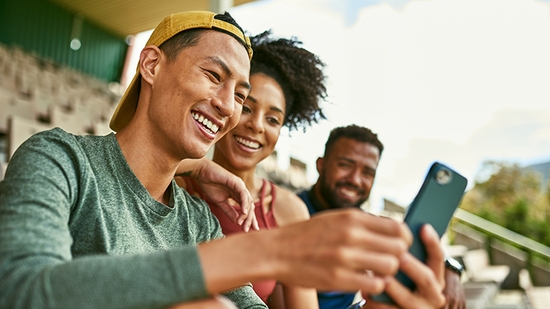 Three happy friends looking at a smartphone while sitting outdoors.