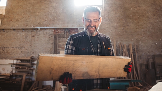A smiling carpenter wearing safety glasses and gloves holds a large piece of raw lumber in a sunlit woodworking shop.