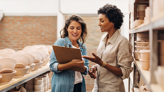 Two women in a pottery studio discuss information on a clipboard while standing next to shelves of ceramic bowls.