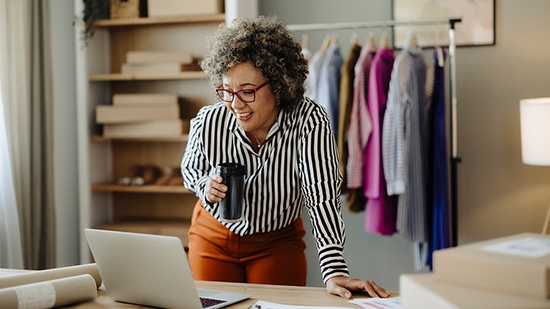 A smiling woman in a striped shirt and red glasses holds a cup while looking at a laptop in a small business setting with clothes racks in the background.