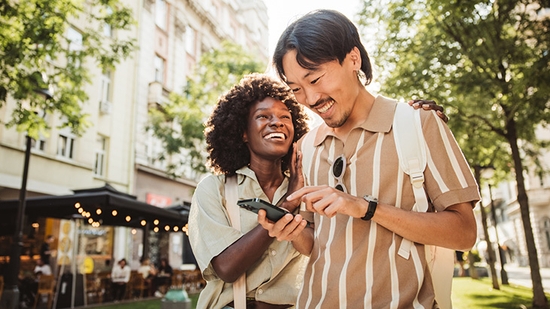 A happy young couple stands on a sunny city street looking at a smartphone together.