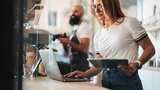 A woman in glasses and a white T-shirt works on a laptop at a cafe counter while holding a clipboard, with a barista visible in the background.