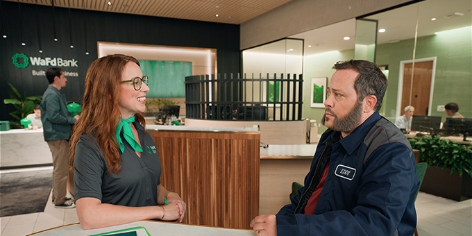 A smiling WaFd Bank employee wearing a green scarf talks to a male customer in a work jacket inside a WaFd branch.