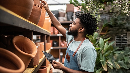 A male business owner in an apron uses a tablet while inspecting terracotta pots on a shelf in a lush plant nursery.