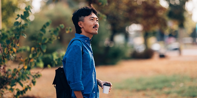 A man in a blue shirt holding a coffee cup while standing outdoors in a park setting.