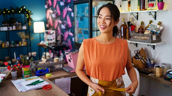 A smiling small business owner, wearing an apron, stands behind the counter of her retail store.