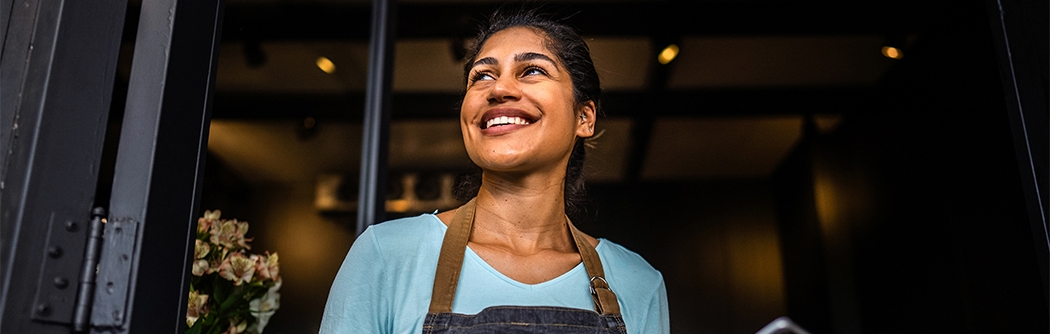 Smiling small business owner in an apron holds a tablet while standing in the doorway of her shop.