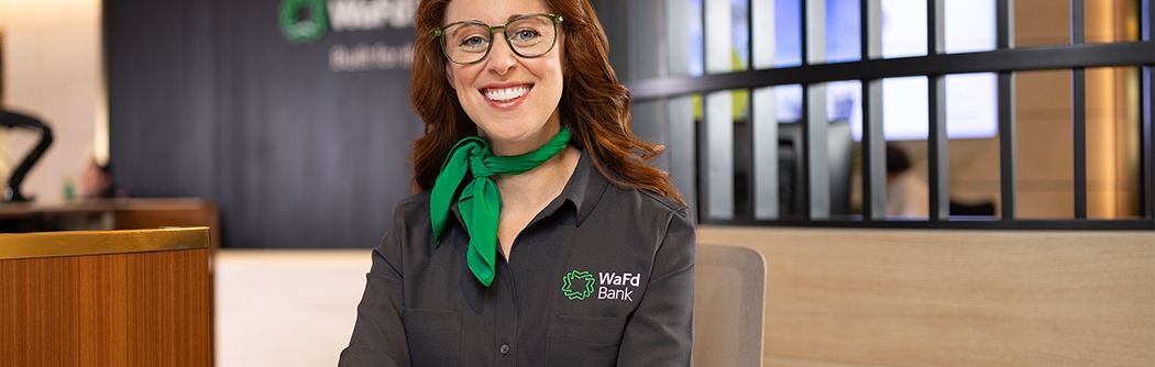A smiling WaFd Bank representative in a branded shirt and green scarf sits at a desk, providing professional support to business clients.
