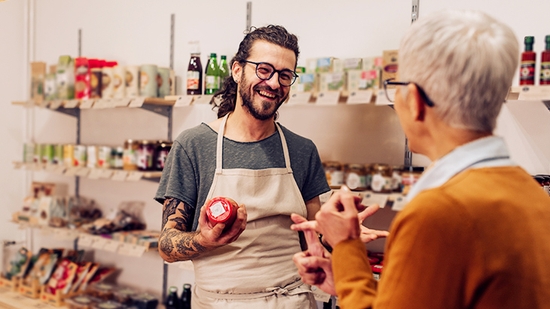 A smiling grocer in an apron talks to a customer in his store, holding up a jar of food.