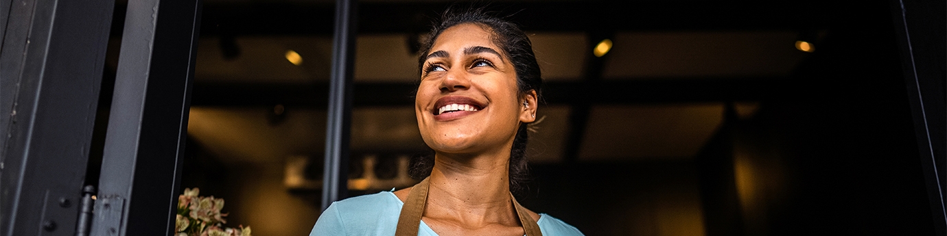 Smiling small business owner in an apron holds a tablet while standing in the doorway of her shop.
