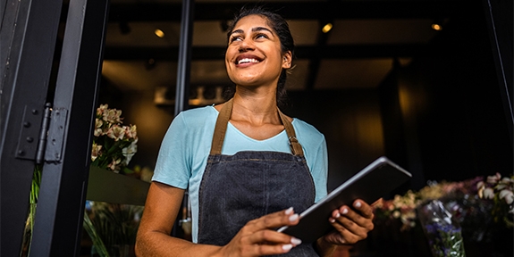 Smiling small business owner in an apron holds a tablet while standing in the doorway of her shop.