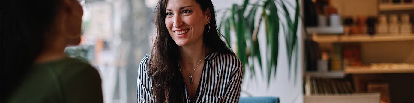 A woman in a striped shirt smiles while holding a tablet and talking to a colleague in a bright, modern cafe setting.