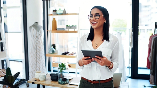 A smiling young small business owner stands in her boutique holding a digital tablet.