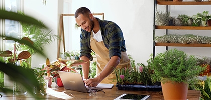A business owner in an apron working on a laptop at a wooden table in a plant-filled shop or nursery.