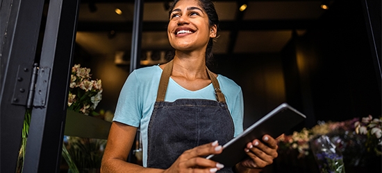 Smiling small business owner in an apron holds a tablet while standing in the doorway of her shop.