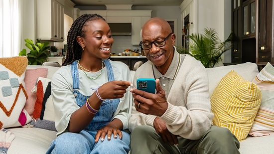 A young woman sits on a couch with an older man, showing him something on her smartphone.