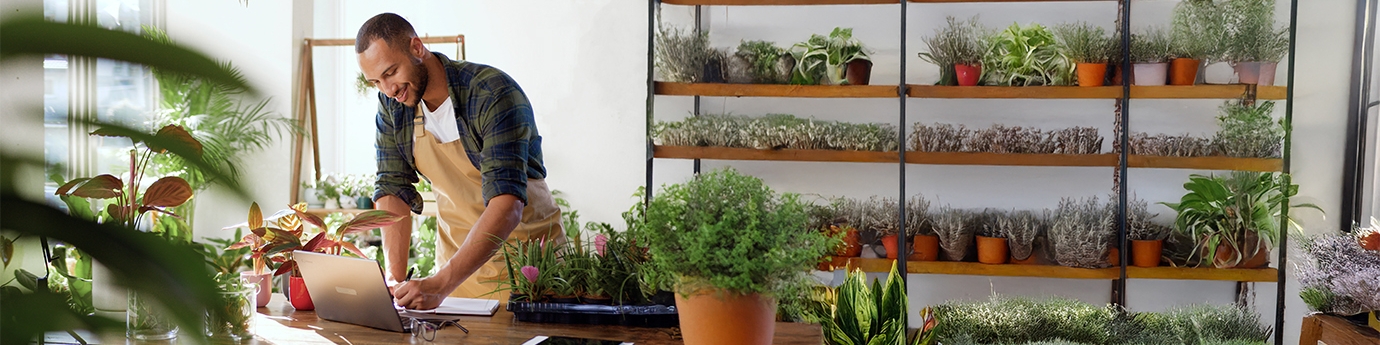 A business owner in an apron working on a laptop at a wooden table in a plant-filled shop or nursery.