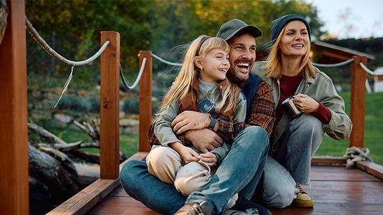 A smiling family of three—a man, woman, and young girl—sit together on a wooden outdoor bridge.