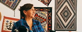 An older woman in a plaid shirt stands in a shop featuring Native American jewelry and woven rugs on the wall.