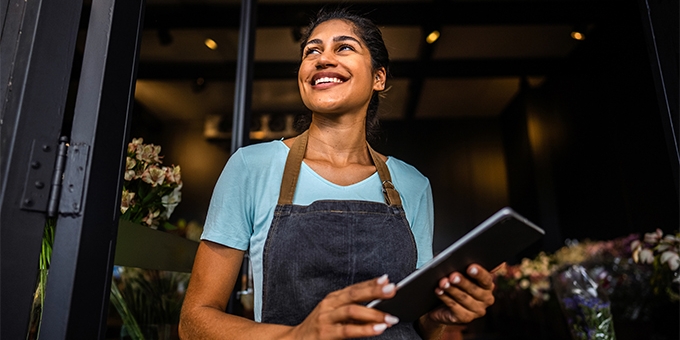 Smiling small business owner in an apron holds a tablet while standing in the doorway of her shop.
